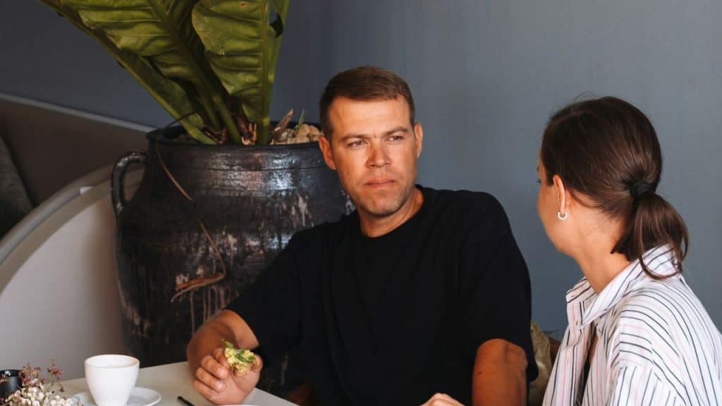 A man in a black t-shirt looks seriously at a woman sitting across the table from him.