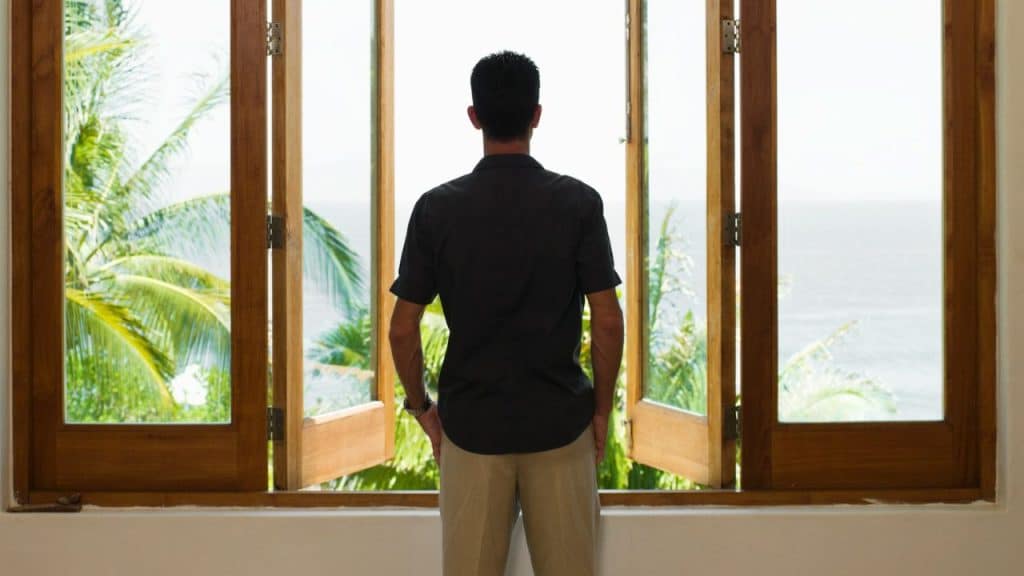 Man standing with his back to the camera looks out open wooden windows at a tropical view.