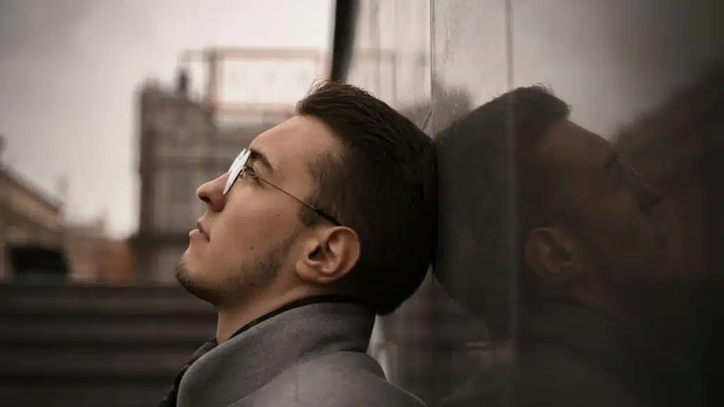 Young man in a gray coat and glasses leans his head on a dark wall outdoors.