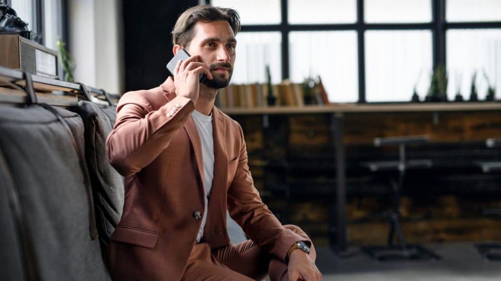 A man in a brown suit sitting indoors while talking on his phone.