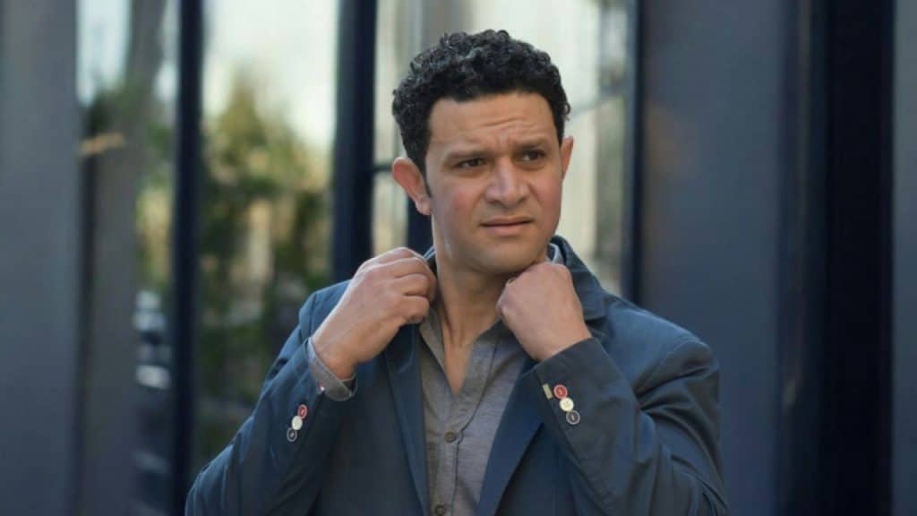 A man with dark, curly hair adjusts the collar of his blue blazer outdoors.