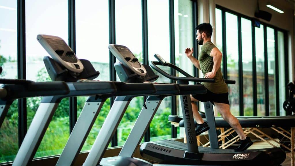 A man running on a treadmill in a gym with large windows.