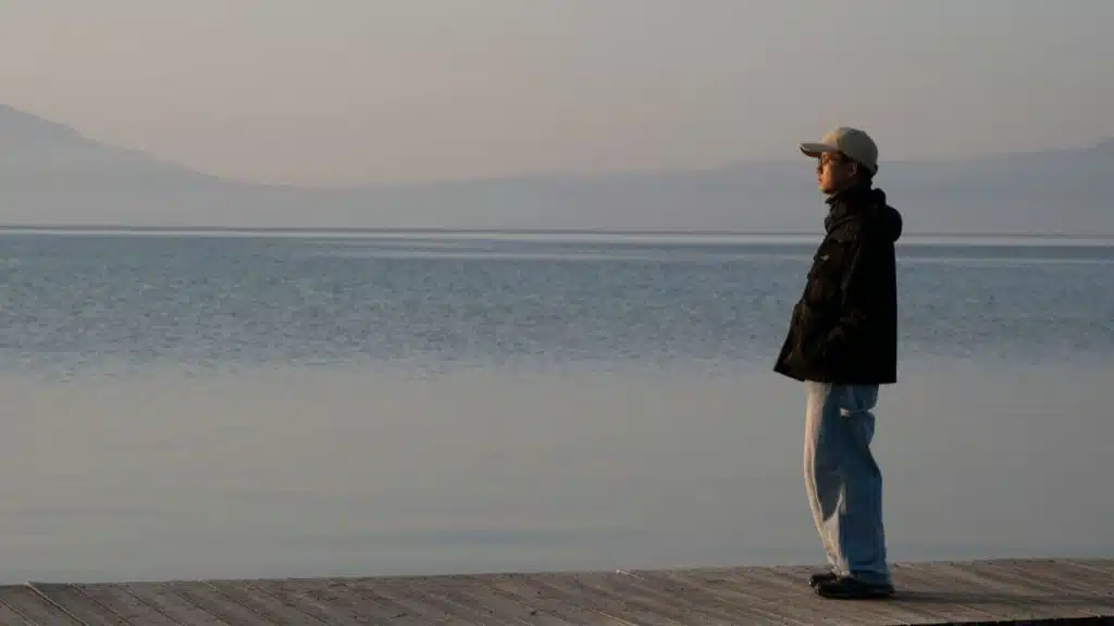 A person in a black jacket, light jeans, and a baseball cap stands on a wooden dock looking out over calm water toward distant, hazy mountains.