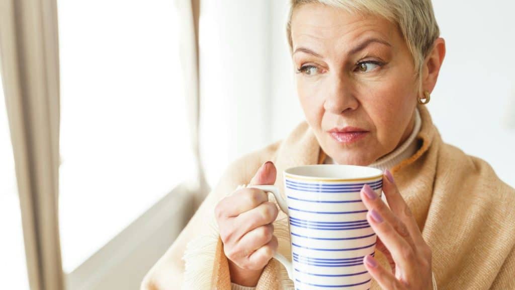 A blonde woman in a tan blanket holds a striped blue and white mug by a bright window.