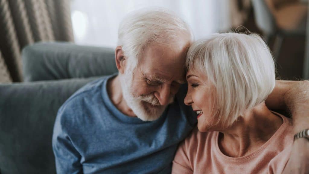 Smiling senior couple embracing on a sofa, forehead to forehead in a gentle moment.