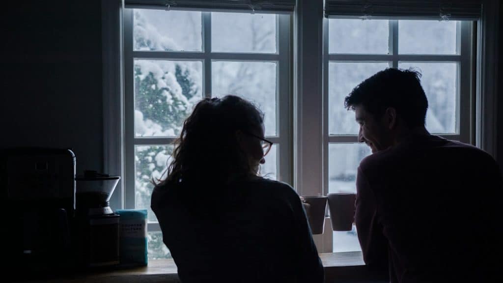 A silhouette of a couple talking over coffee by a window, looking out at a snowy scene.
