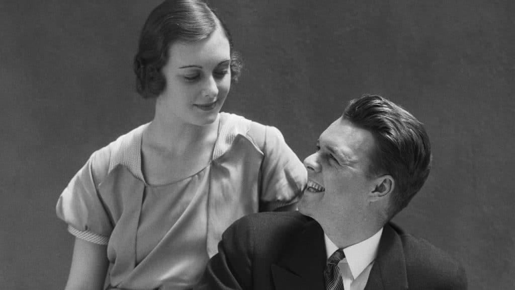 A black and white photo of a man and woman looking at each other and smiling, with the man seated and the woman standing behind him.