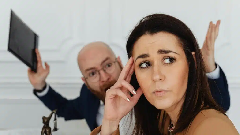 A woman looking stressed while a man in the background gestures angrily.