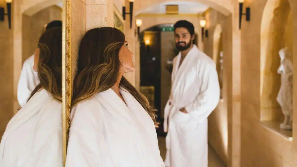 A couple in white bathrobes standing in a warmly lit hallway.