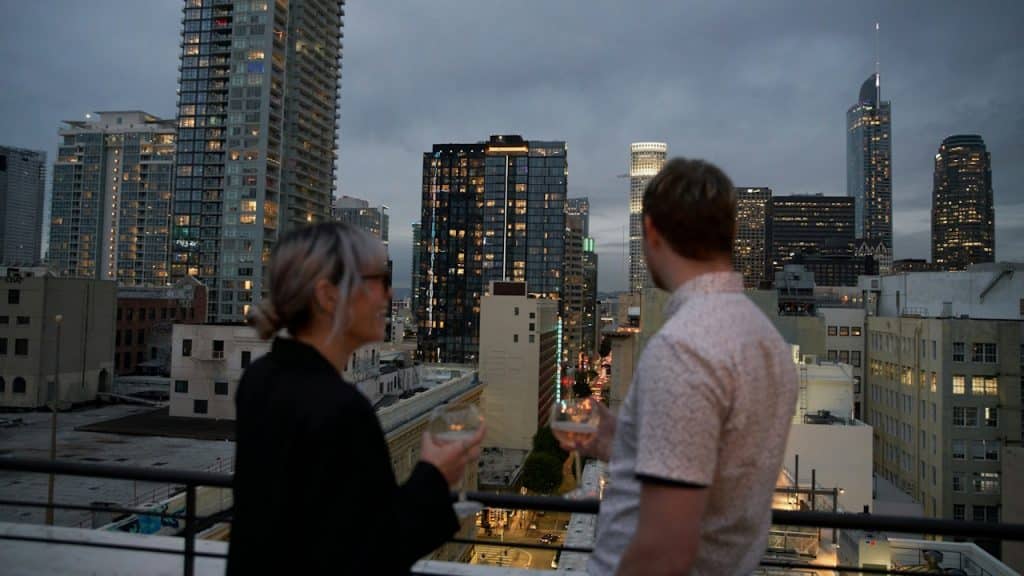 A couple on a rooftop at dusk, holding wine glasses and looking out at the city skyline of tall buildings.