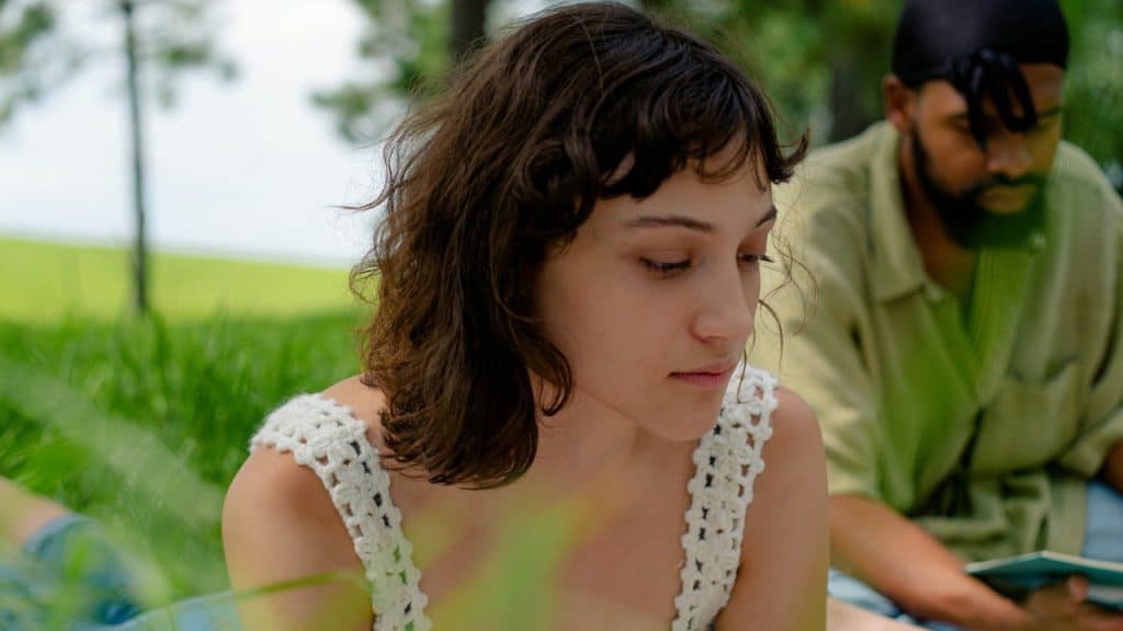 A young woman wearing a white crocheted top, sitting outdoors in the grass with a man in the background.