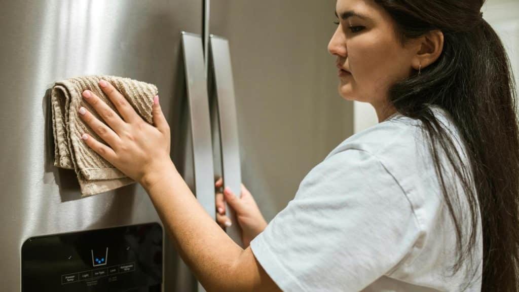 A woman cleaning a stainless steel refrigerator with a cloth.