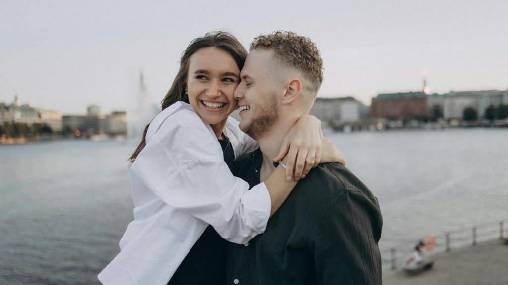 A smiling couple embracing by the water with a cityscape in the background.