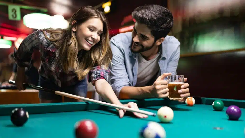 A couple smiling and playing pool together, with one holding a drink.