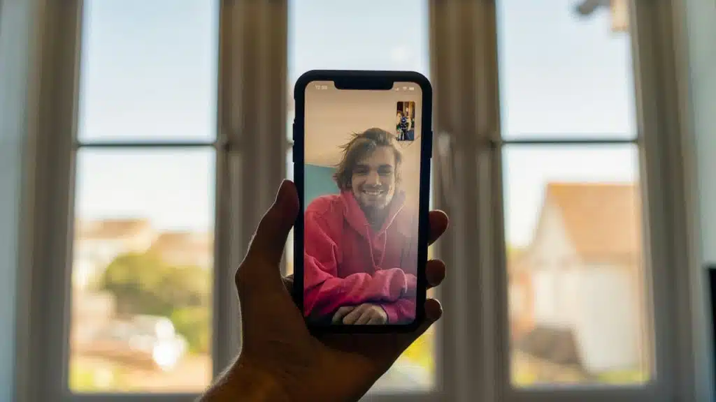 A person holds a smartphone displaying a video call with a smiling man in a pink hoodie on the screen, framed against a window with an outdoor view.
