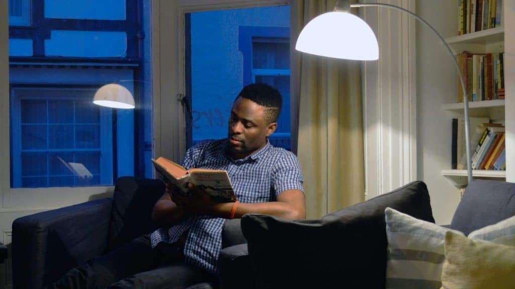 A man sits comfortably on a sofa reading a book under a floor lamp at night.