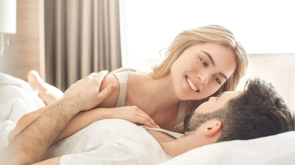 A smiling woman is leaning over a man lying on his back in bed.
