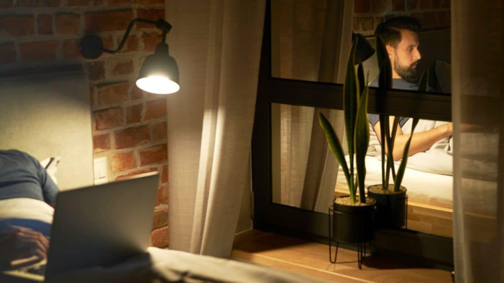 A man is visible through a window in a brick wall in the background, while a laptop is on a bed in the foreground.