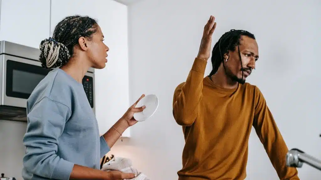A man and a woman are arguing in a kitchen, with the man raising his hand.
