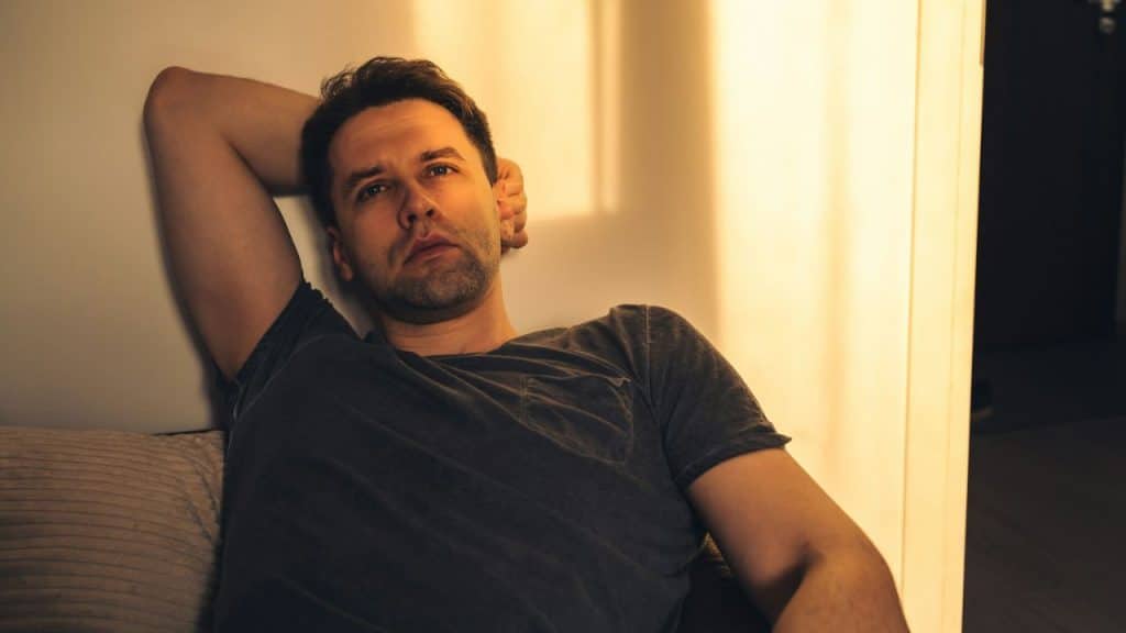 A man in a gray shirt is sitting with his arm behind his head, looking forward.