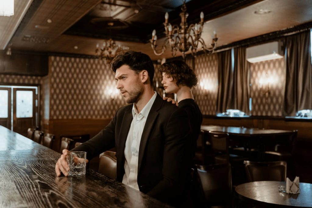 Man in Black Suit Sitting on a Bar Holding a Glass of Whiskey Beside a Woman in Black Dress