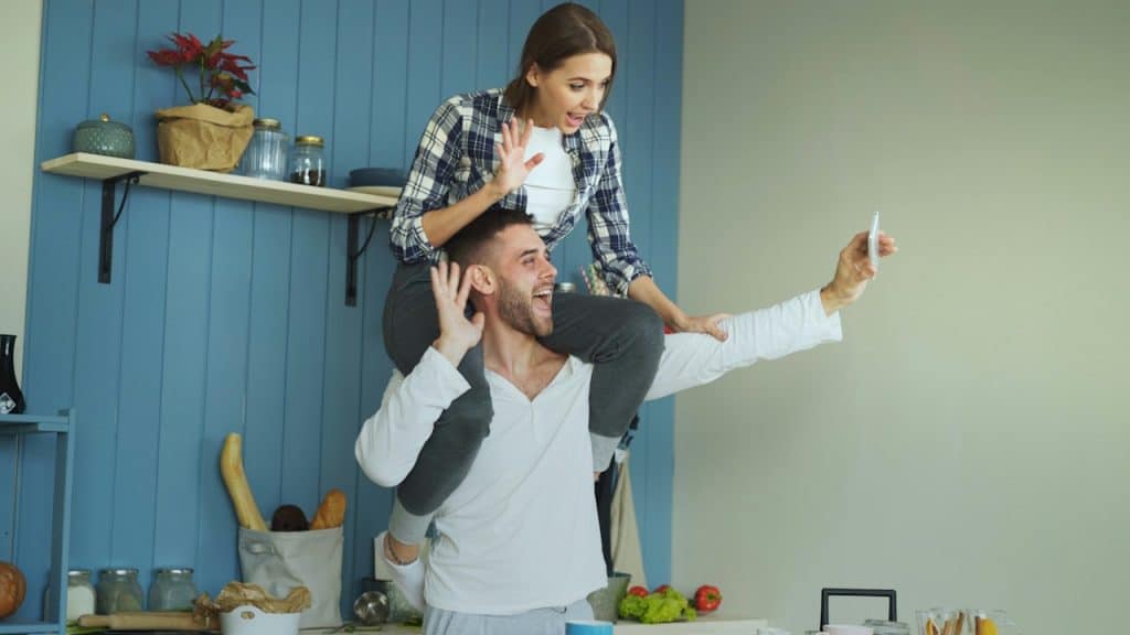 A laughing man gives a woman a piggyback ride and holds up a smartphone to take a selfie in a kitchen.