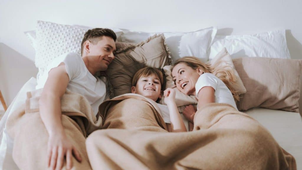 A mother, father, and child are smiling while lying in a bed with blankets.