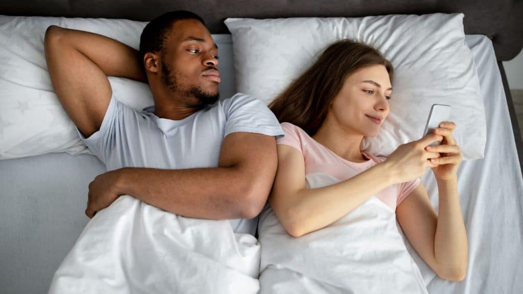 An overhead shot of a Black man and a White woman lying in bed; the woman uses her phone while the man looks at her.