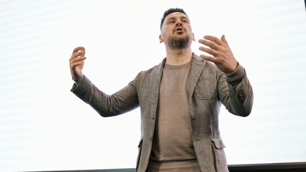 A man with a beard and gray blazer gestures with his hands while speaking.
