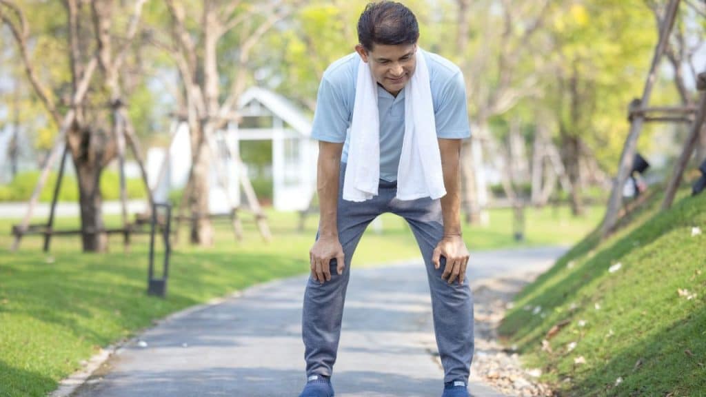 A smiling, middle-aged man is catching his breath after a run in a sunny park.