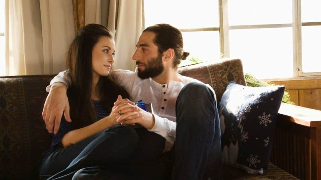 Couple seated on a couch by a window, looking at each other and holding hands.