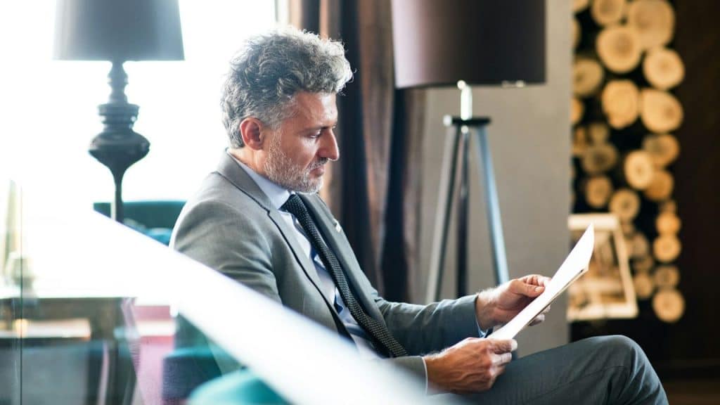 Well-dressed man with gray hair and a beard, sitting in an armchair and reading a document.
