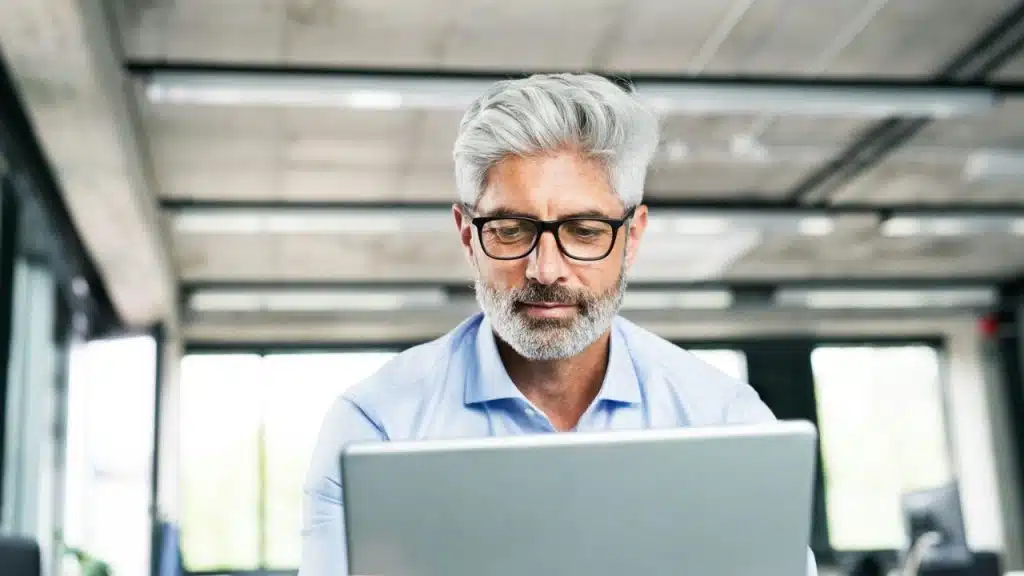 Man with gray hair and beard wearing glasses looking at a laptop.