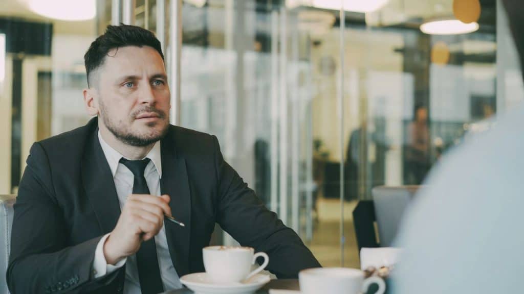 Man in a suit and tie sits at a table with coffee cups, looking intently at someone.