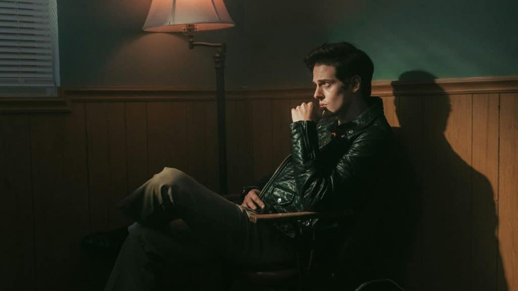 A man in a leather jacket sits in a chair, thoughtfully looking away in a dimly lit room.