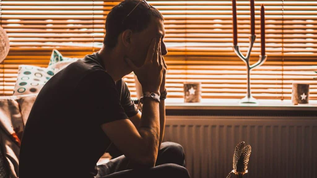 A man sits in profile, hands on his face, next to a window with wooden blinds.