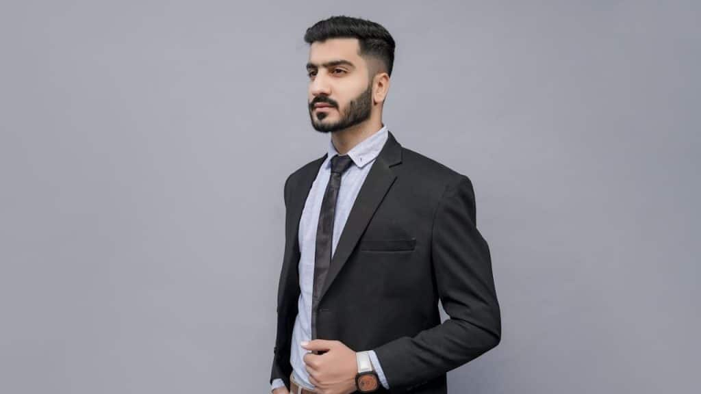 Man in a black suit and tie, with a beard and dark hair, posing against a gray background.