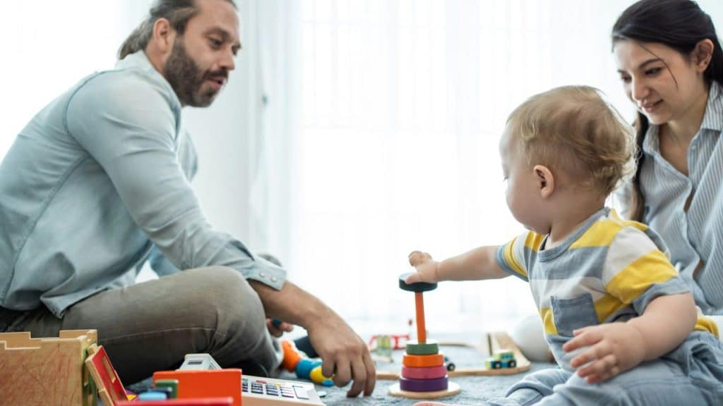 A man and woman sit on the floor watching a baby play with a colorful stacking toy.