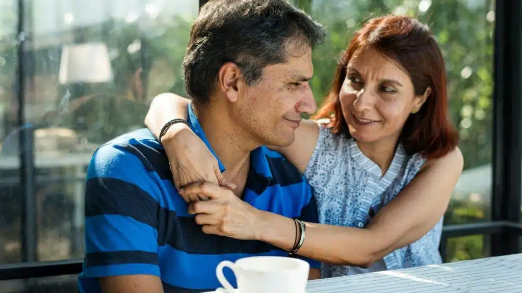 A middle-aged man and woman embrace and smile at each other at a table outdoors.