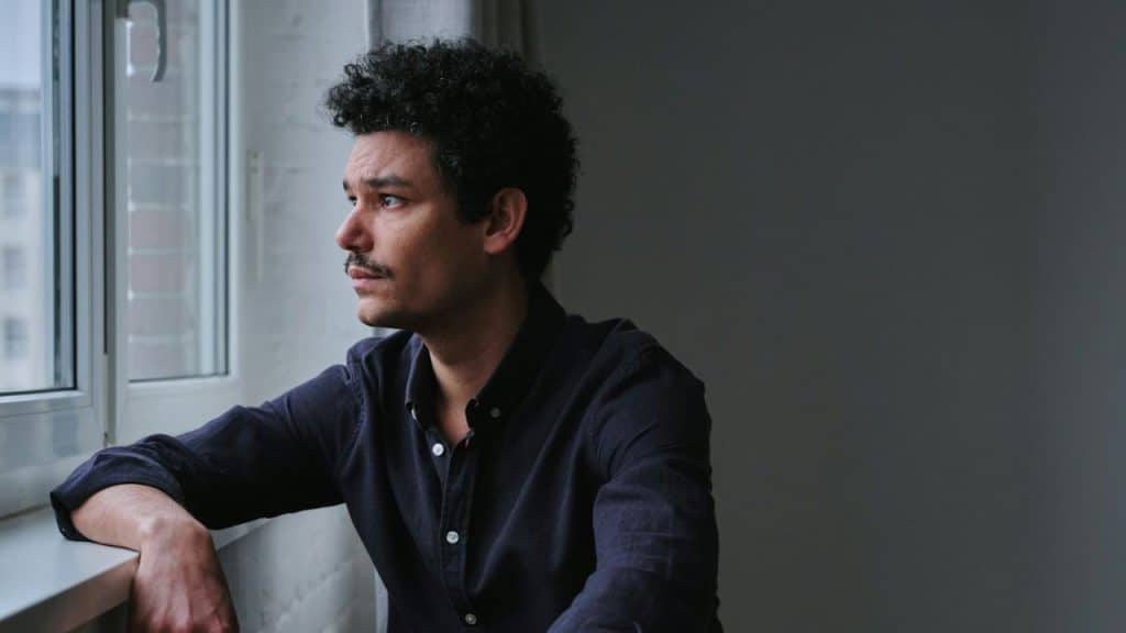 Man with a mustache and curly hair in a dark shirt looking out a window.