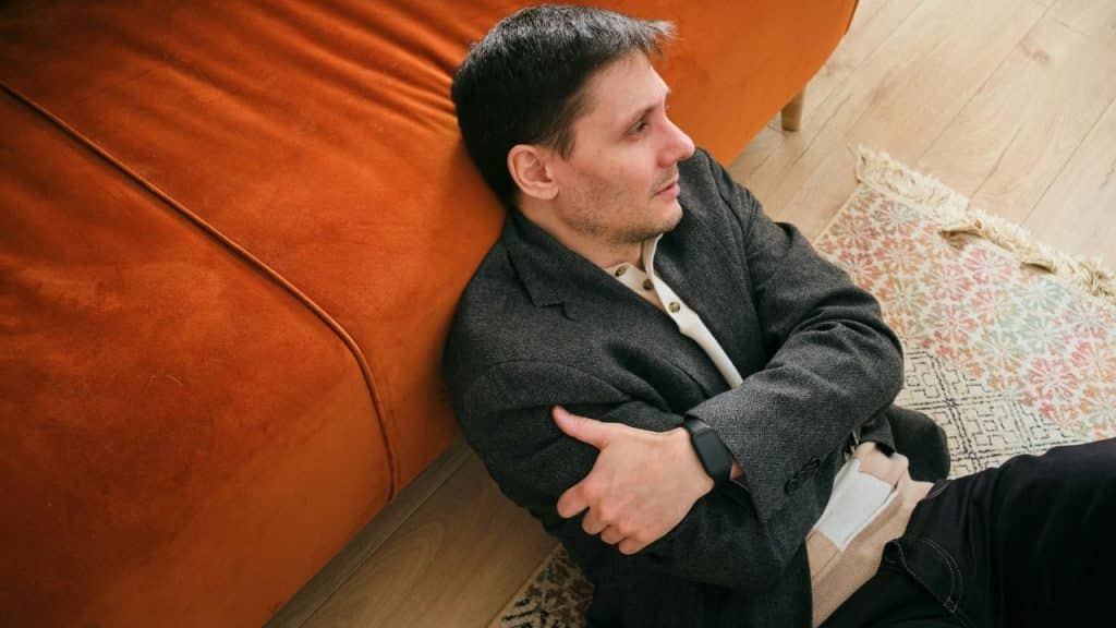A man sits on the floor, leaning against an orange couch, with his arms crossed tightly.