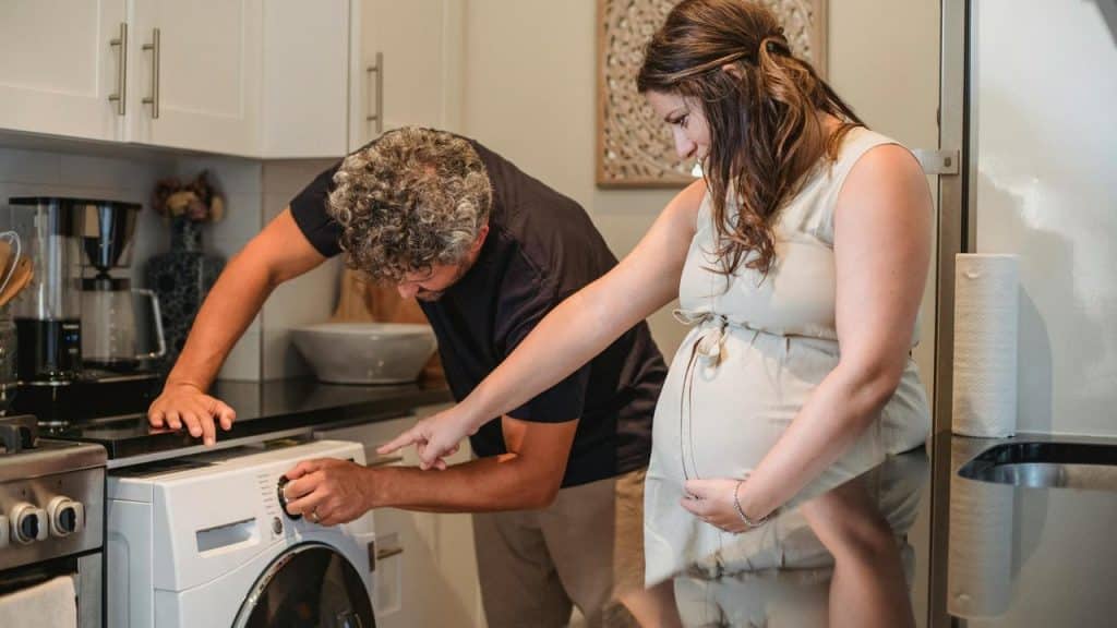 A pregnant woman and a man adjusting settings on a washing machine together.
