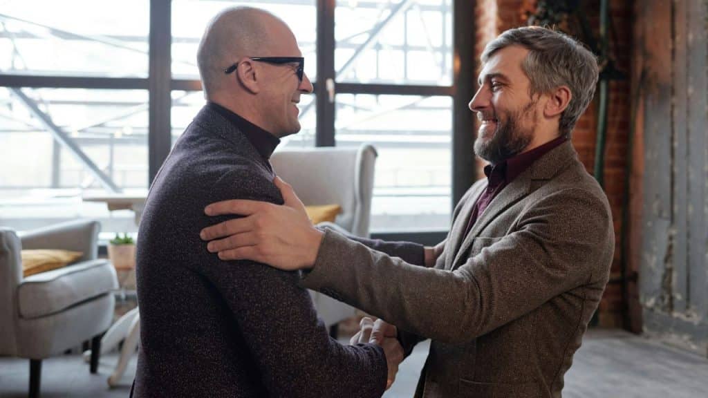 Two businessmen are shaking hands and smiling warmly while embracing indoors.