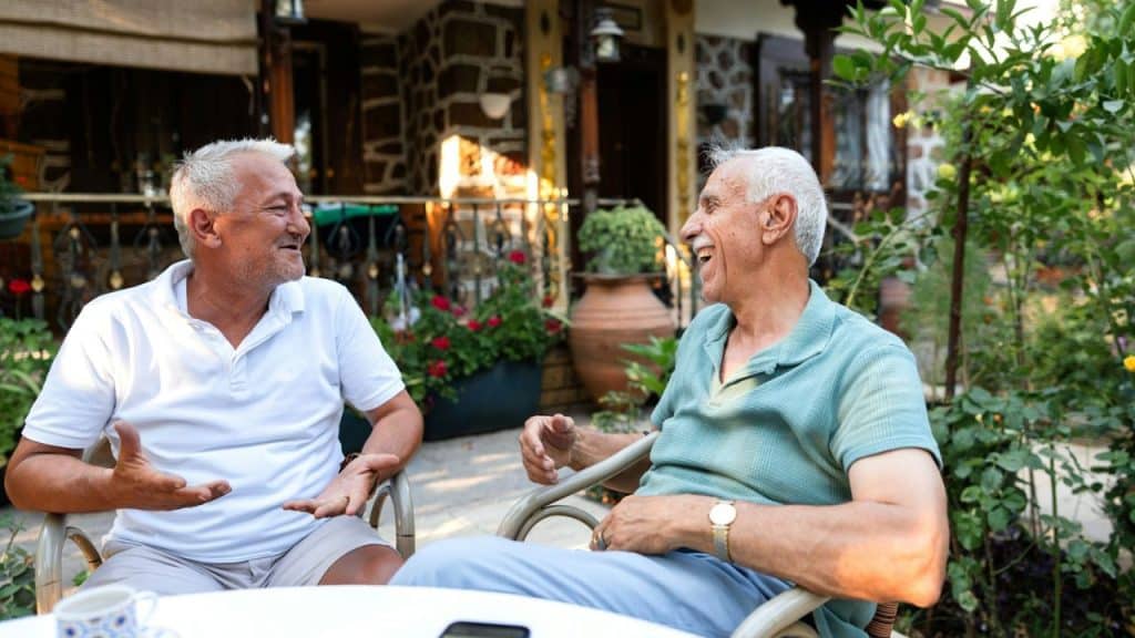 Two older men laughing while sitting in chairs outdoors on a sunny patio.