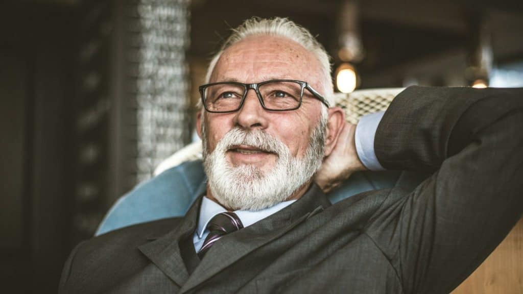 Mature man with white beard, glasses, and suit, leaning back, looking up.