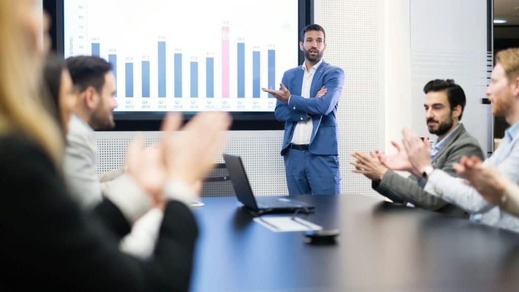 Man presenting data charts to colleagues who are seated and applauding him.