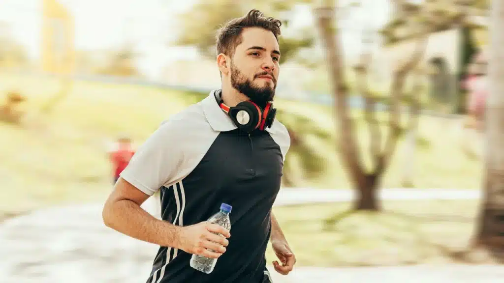 Bearded man with headphones around his neck running while holding a water bottle.