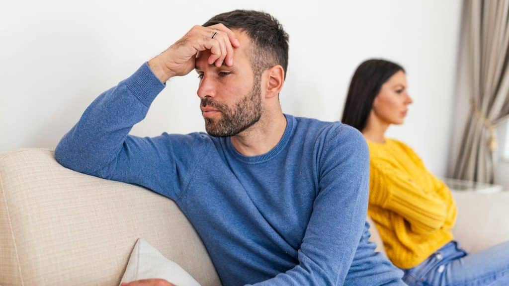 A distraught man in a blue shirt sits on a couch, facing away from an angry woman with crossed arms.