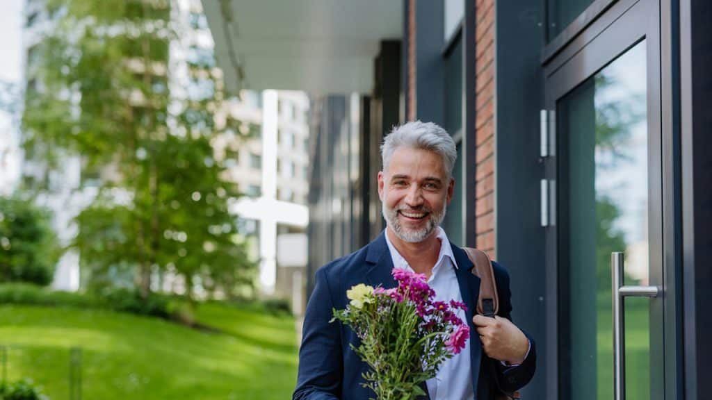 A middle-aged man with gray hair and a beard looks happily at the camera while holding flowers, standing near a glass doorway.