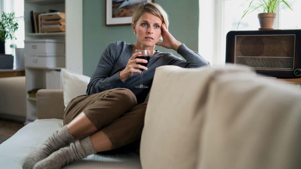 A woman on a couch holds a glass of red wine, looking away with a pensive expression.